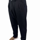 A person wearing black pleated trousers with tan suede shoes, shown from the waist down against a plain white background.
