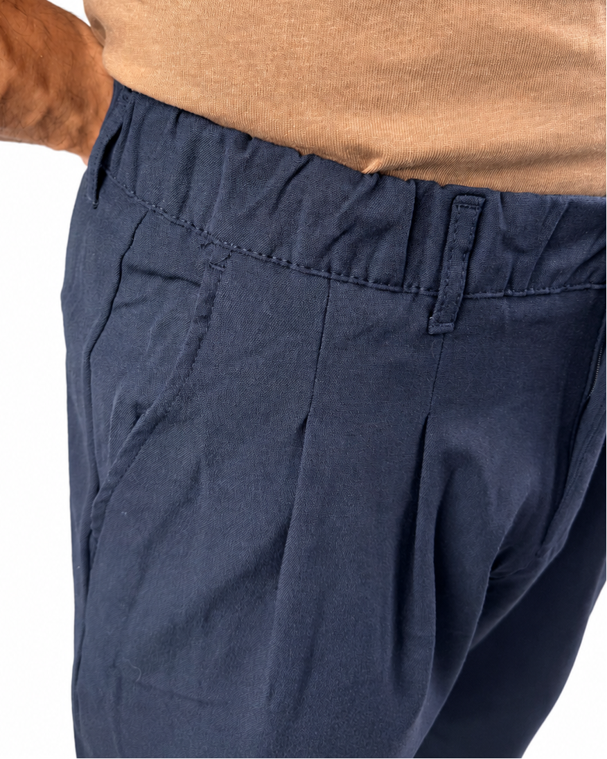 Close-up of a person wearing navy blue pleated trousers and a tan t-shirt, focusing on the waistband and side pocket details against a white background.