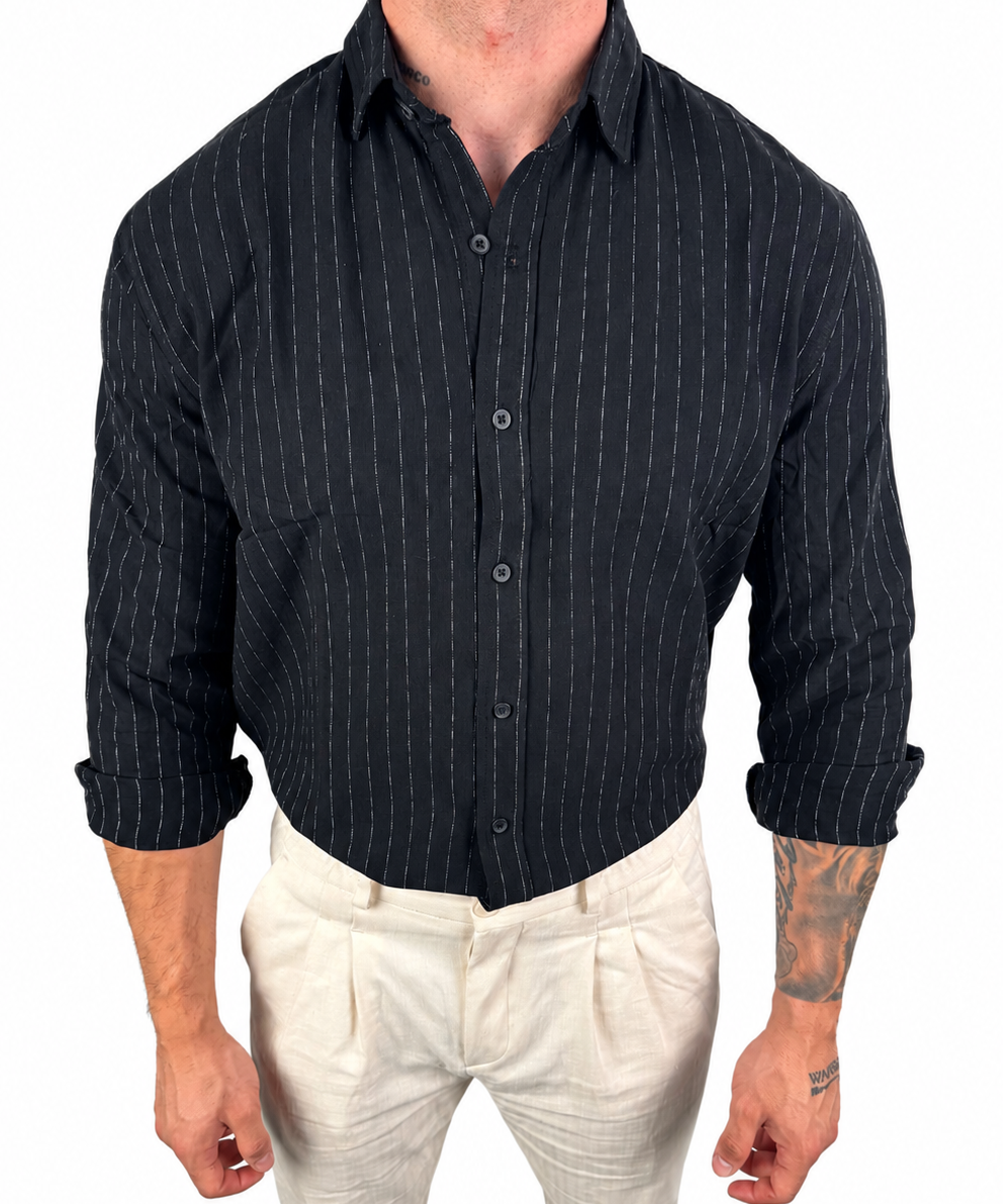 A man wearing a black long-sleeve button-down shirt with thin white vertical pinstripes and light beige trousers, shown from the neck down against a plain white background.
