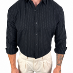 A man wearing a black long-sleeve button-down shirt with thin white vertical pinstripes and light beige trousers, shown from the neck down against a plain white background.