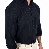 A man wearing a dark navy blue long-sleeve button-down shirt with thin vertical white pinstripes, paired with light beige trousers, shown from the waist down against a plain white background.