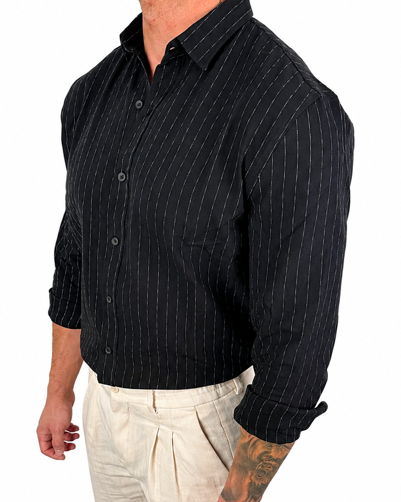 A man wearing a black long-sleeve button-up shirt with thin white vertical pinstripes and light beige trousers, shown from the waist up against a plain white background.