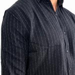 A man wearing a dark navy blue long-sleeve button-down shirt with thin white vertical pinstripes, paired with off-white trousers, shown from the waist up against a plain white background.