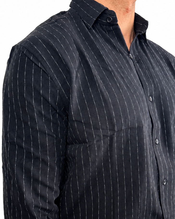 A man wearing a dark navy blue long-sleeve button-down shirt with thin white vertical pinstripes, paired with off-white trousers, shown from the waist up against a plain white background.