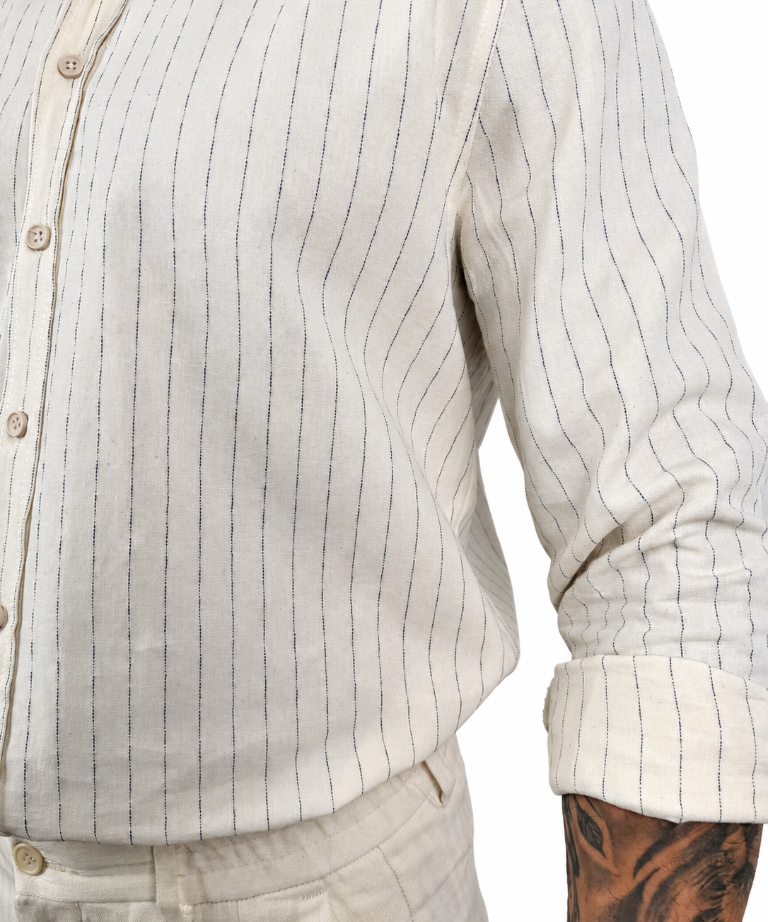 Close-up of a person wearing a light beige cotton button-up shirt with thin dark vertical pinstripes and rolled sleeves, paired with matching beige trousers. A tattooed arm is visible at the side.