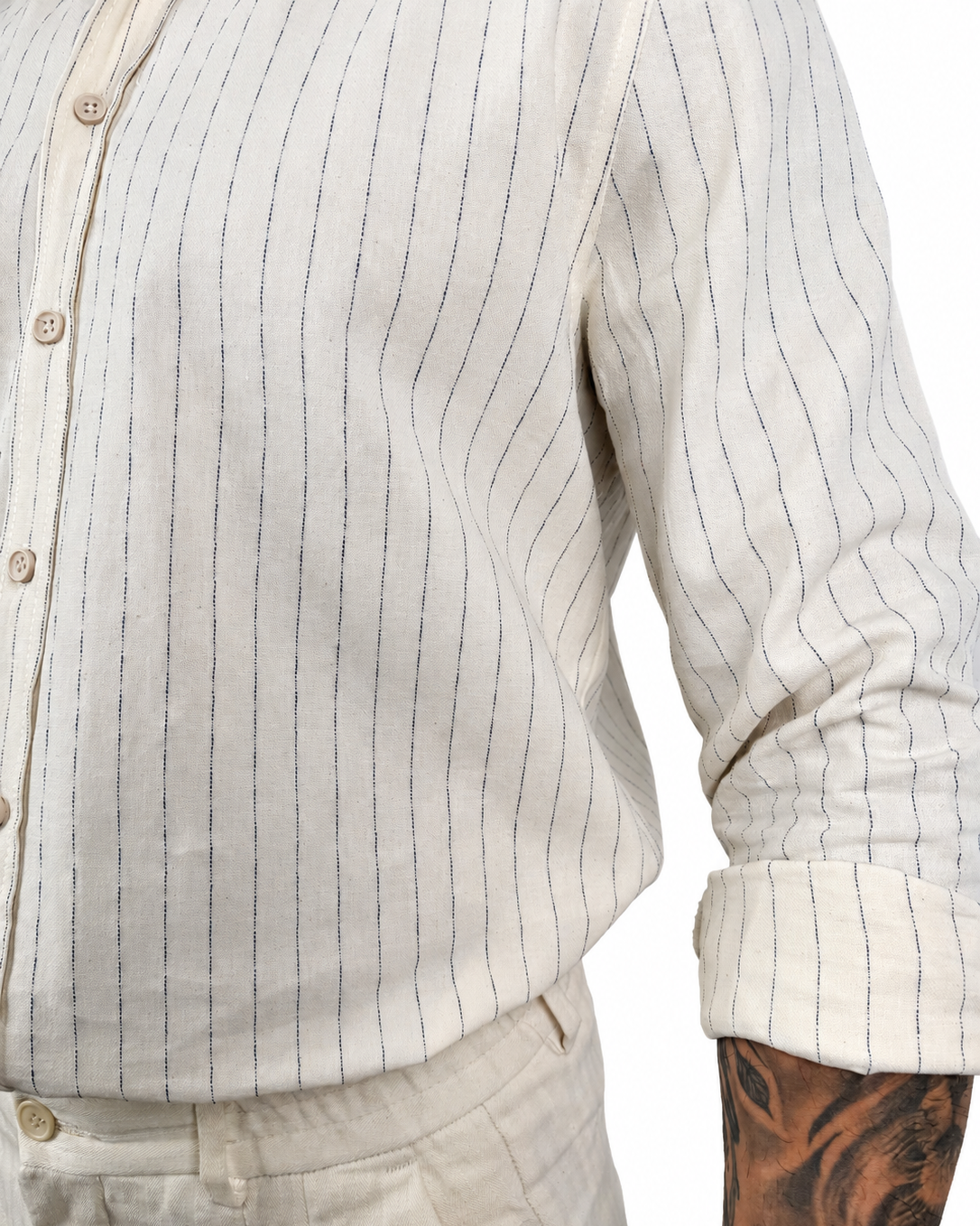 Close-up of a person wearing a light beige cotton button-up shirt with thin dark vertical pinstripes and rolled sleeves, paired with matching beige trousers. A tattooed arm is visible at the side.