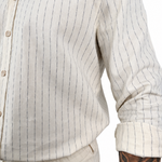 Close-up of a person wearing a light beige cotton button-up shirt with thin dark vertical pinstripes and rolled sleeves, paired with matching beige trousers. A tattooed arm is visible at the side.