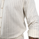 Close-up of a person wearing a light beige cotton button-up shirt with thin dark vertical pinstripes and rolled sleeves, paired with matching beige trousers. A tattooed arm is visible at the side.