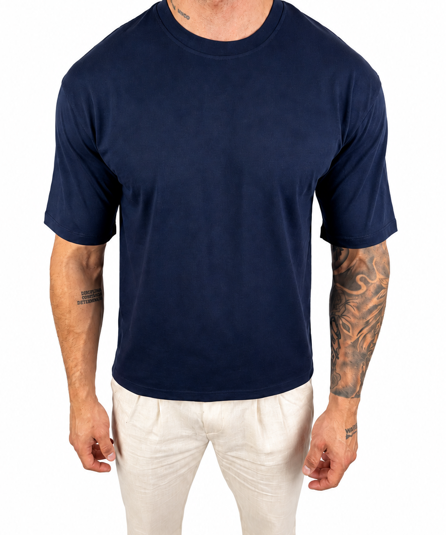 A man wearing a plain navy blue short-sleeve crewneck t-shirt and off-white pants, standing against a solid white background.