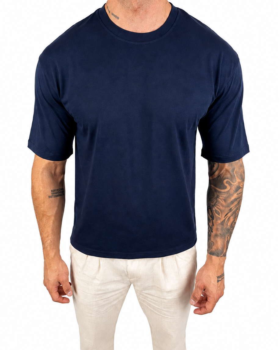 A man wearing a plain navy blue short-sleeve crewneck t-shirt and off-white pants, standing against a solid white background.