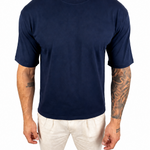 A man wearing a plain navy blue short-sleeve crewneck t-shirt and off-white pants, standing against a solid white background.