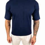 A man wearing a plain navy blue short-sleeve crewneck t-shirt and off-white pants, standing against a solid white background.