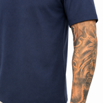 A close-up of a person wearing a navy blue short-sleeve t-shirt and off-white trousers, showing a detailed tattoo on the arm.