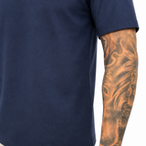 A close-up of a person wearing a navy blue short-sleeve t-shirt and off-white trousers, showing a detailed tattoo on the arm.