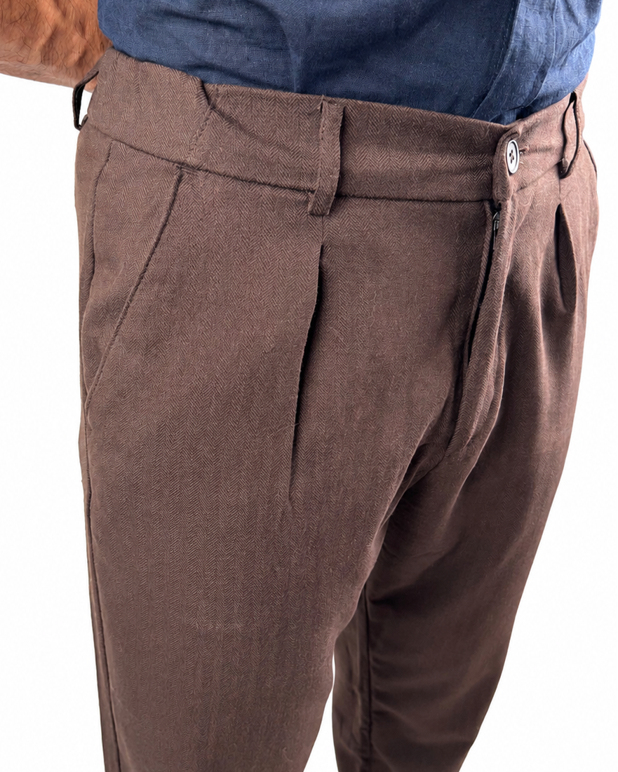 A close-up of a person wearing brown pleated trousers and a blue button-up shirt against a white background.
