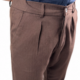 A close-up of a person wearing brown pleated trousers and a blue button-up shirt against a white background.