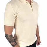 A man wearing a cream-colored short-sleeve polo shirt with a subtle grid texture, paired with black pants, shown from the side against a plain white background.