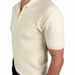A man wearing a short-sleeve knit polo shirt in cream with a subtle grid texture, paired with black pants, shown from a side profile against a white background.