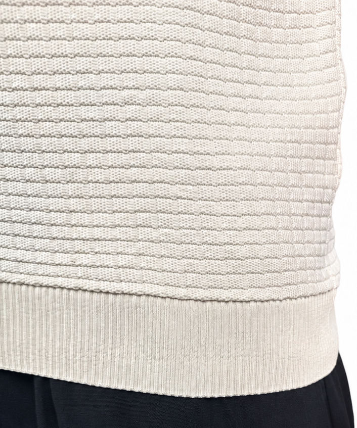 Close-up of a person wearing a cream ribbed knit sweater vest and black pants against a white background.