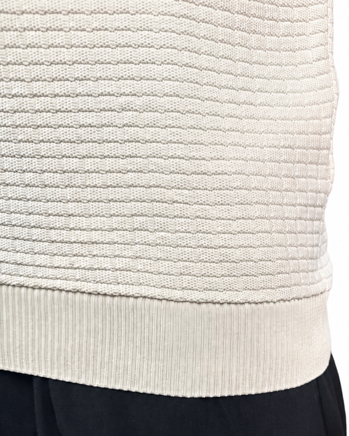Close-up of a person wearing a cream ribbed knit sweater vest and black pants against a white background.
