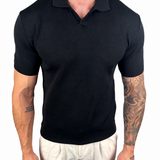 A man wearing a black short-sleeve polo shirt and off-white trousers, shown from the neck down against a plain white background.