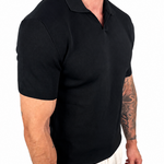 A man wearing a black short-sleeve polo shirt and light beige trousers, shown from the side against a plain white background.