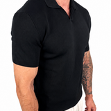 A man wearing a black short-sleeve polo shirt and light beige trousers, shown from the side against a plain white background.
