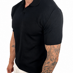 A man wearing a black short-sleeve polo shirt and off-white trousers, shown from the side against a plain white background.
