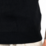 Close-up of a person wearing a black ribbed knit polo and light beige linen trousers, with the right arm visible showing a large tattoo and a small 'WARRIOR' wrist tattoo.