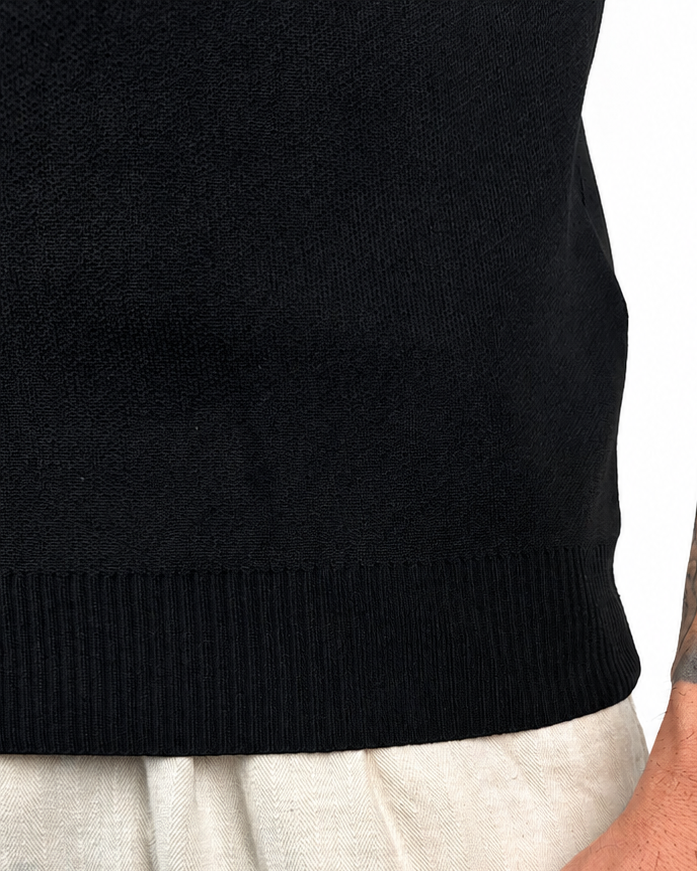 Close-up of a person wearing a black ribbed knit polo and light beige linen trousers, with the right arm visible showing a large tattoo and a small 'WARRIOR' wrist tattoo.
