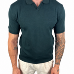 A man wearing a dark teal short-sleeve polo shirt and off-white trousers, shown from the neck down against a plain white background.