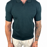 A man wearing a dark teal short-sleeve polo shirt and off-white trousers, shown from the neck down against a plain white background.