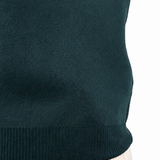 Close-up of a person wearing a dark teal ribbed knit sweater and light beige trousers, with a tattooed arm visible against a plain white background.