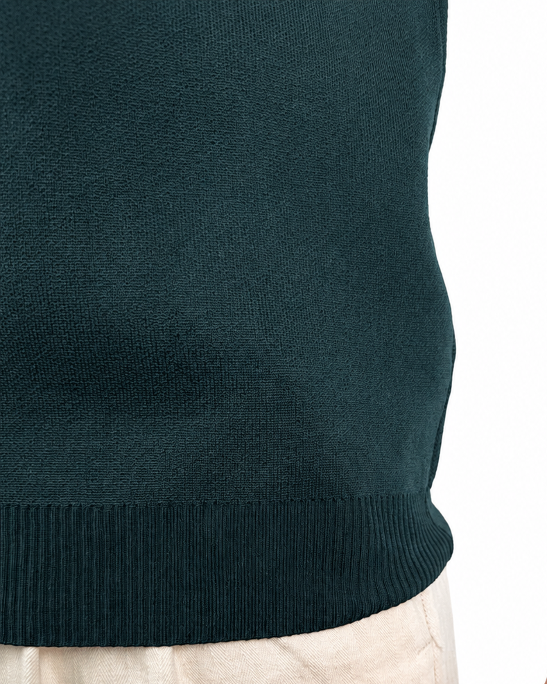 Close-up of a person wearing a dark teal ribbed knit sweater and light beige trousers, with a tattooed arm visible against a plain white background.