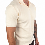 A man wearing a cream-colored short-sleeve knit polo shirt and dark brown trousers, shown from a side profile against a plain white background.