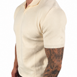 A man wearing a cream-colored short-sleeve knit polo shirt, dark brown pants, and white socks, shown from a side profile against a plain white background.