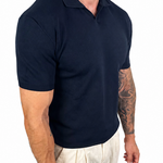 A muscular man wearing a dark navy blue short-sleeve polo shirt and off-white linen trousers, shown from the waist down against a solid white background.
