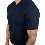 A man wearing a dark navy blue short-sleeve polo shirt and off-white trousers, shown from a side profile against a plain white background.