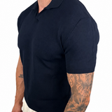 A man wearing a dark navy blue short-sleeve polo shirt and off-white trousers, shown from a side profile against a plain white background.