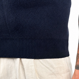 Close-up of a person wearing a dark navy ribbed knit sweater and light beige trousers, with a detailed arm tattoo and a wrist tattoo reading 'WARRIOR' visible against a white background.