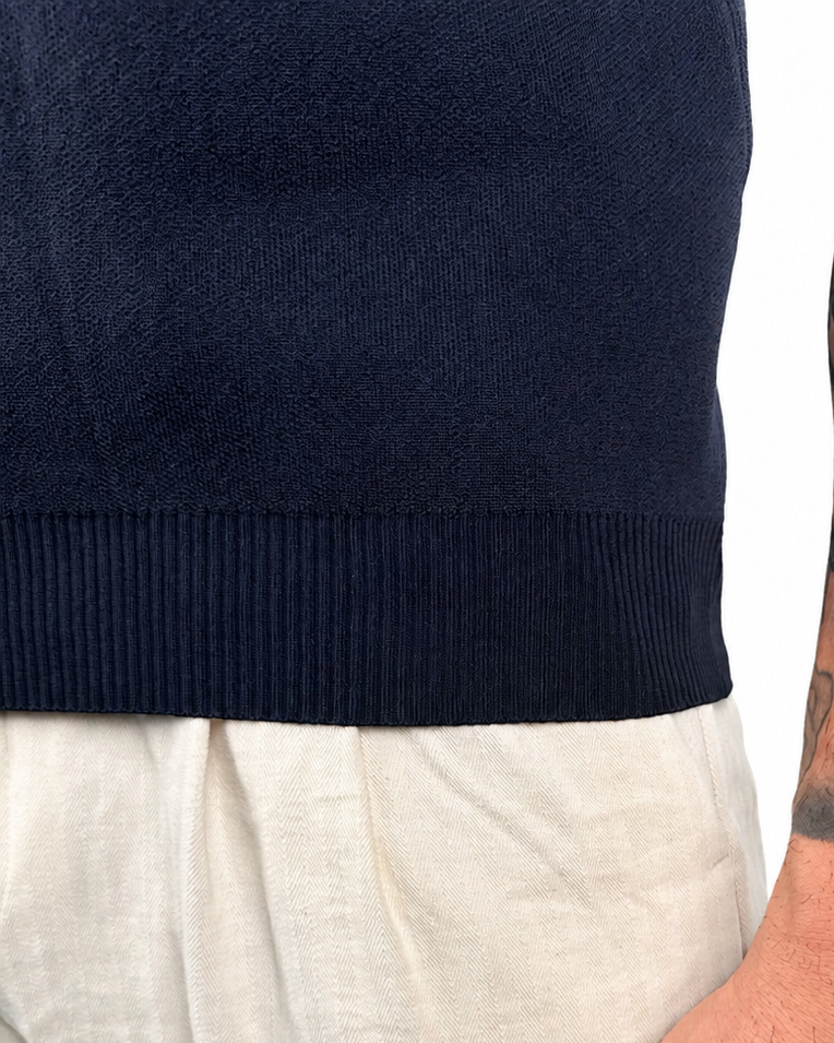 Close-up of a person wearing a dark navy ribbed knit sweater and light beige trousers, with a detailed arm tattoo and a wrist tattoo reading 'WARRIOR' visible against a white background.