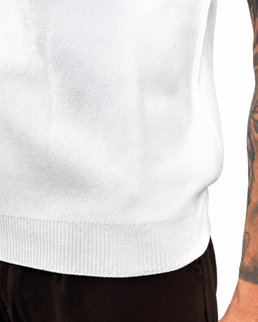 Close-up of a person wearing a white ribbed knit sweater and dark brown pants, with a heavily tattooed arm visible against a plain white background.