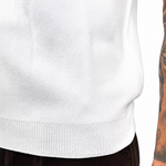 Close-up of a person wearing a white ribbed knit sweater and dark brown pants, with a heavily tattooed arm visible against a plain white background.