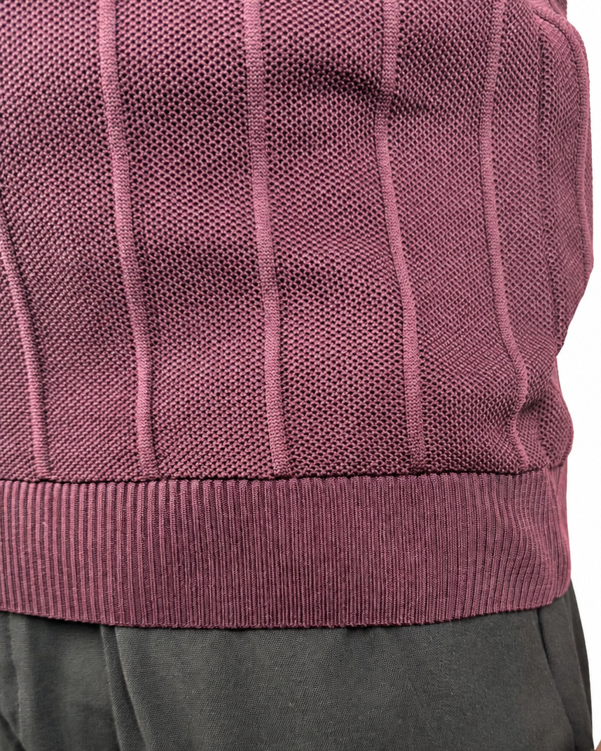 Close-up of a person wearing a ribbed knit sweater vest in a muted mauve-purple color, paired with dark gray trousers, against a plain white background.