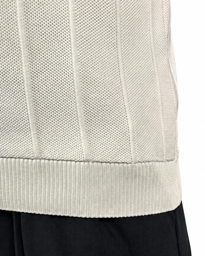 Close-up of a person wearing a cream ribbed knit sweater vest and black pants against a white background.