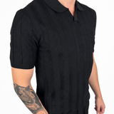 A man wearing a black short-sleeve polo shirt with a textured knit pattern and black pants, shown from the neck down against a plain white background.