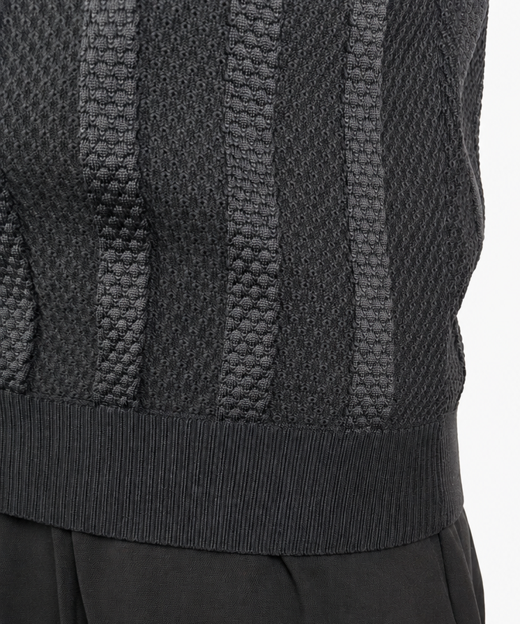 Close-up of a person wearing a dark charcoal gray knit sweater vest with vertical textured stripes and black trousers, against a plain white background.