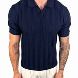 A man wearing a navy blue short-sleeve polo shirt with a textured knit pattern and white pants, shown from the neck down against a plain white background.