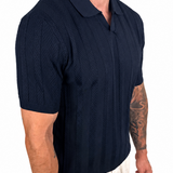A man wearing a dark navy blue short-sleeve polo shirt with a textured knit pattern and light beige trousers, shown from a side profile against a white background.