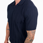A man wearing a dark navy blue short-sleeve knit polo shirt and light beige pants, shown from the waist up against a plain white background.